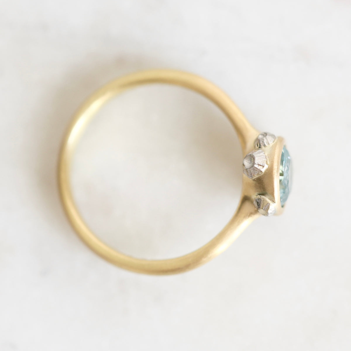 A gold ring with a small cluster of clear stones and a light blue-green gemstone, photographed from above on a light marble-like surface.