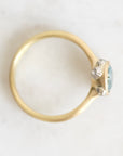 A gold ring with a small cluster of clear stones and a light blue-green gemstone, photographed from above on a light marble-like surface.