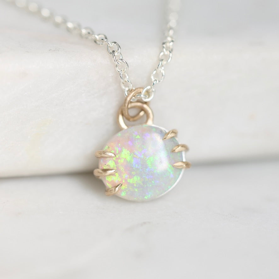 A delicate silver chain necklace with a small oval pendant featuring a white iridescent opal-like stone held in gold-toned prong settings, displayed on a soft white surface.
