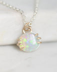 A delicate silver chain necklace with a small oval pendant featuring a white iridescent opal-like stone held in gold-toned prong settings, displayed on a soft white surface.