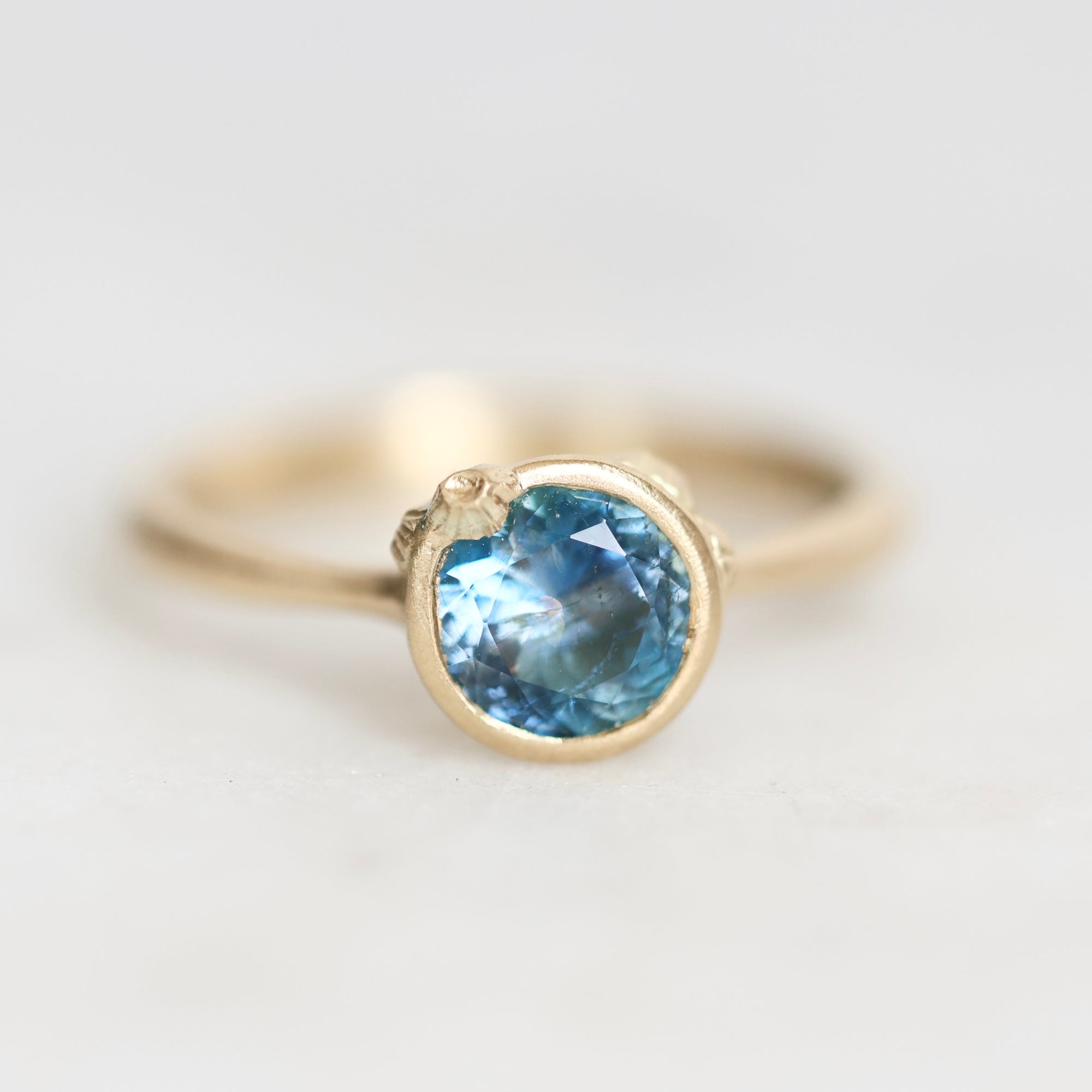 A gold ring featuring a round-cut light blue gemstone in a bezel setting, photographed on a clean white background.