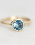 A gold ring featuring a round-cut light blue gemstone in a bezel setting, photographed on a clean white background.