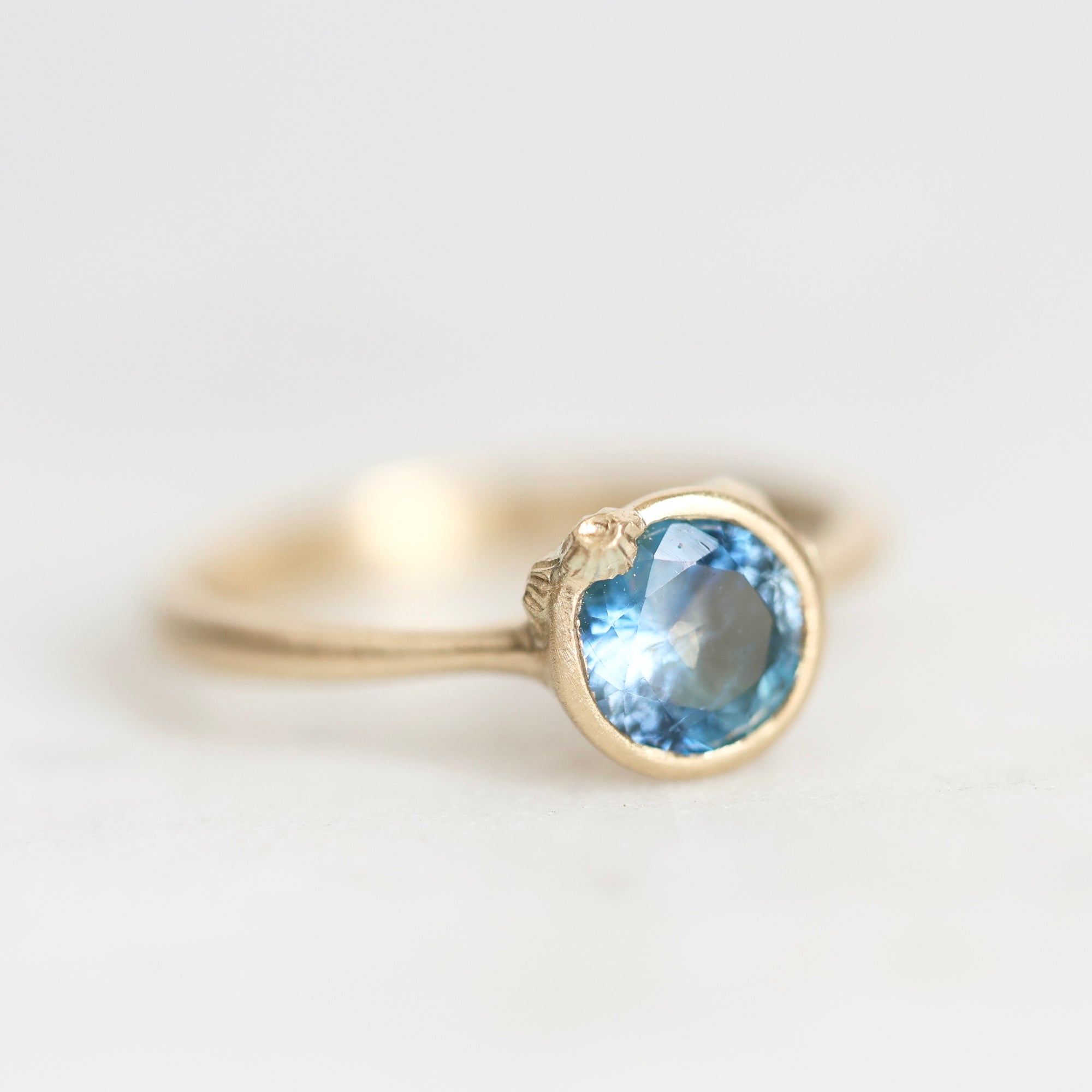 A gold ring featuring a round-cut light blue gemstone in a bezel setting, photographed on a clean, light gray background.