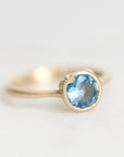 A gold ring featuring a round-cut light blue gemstone in a bezel setting, photographed on a clean, light gray background.