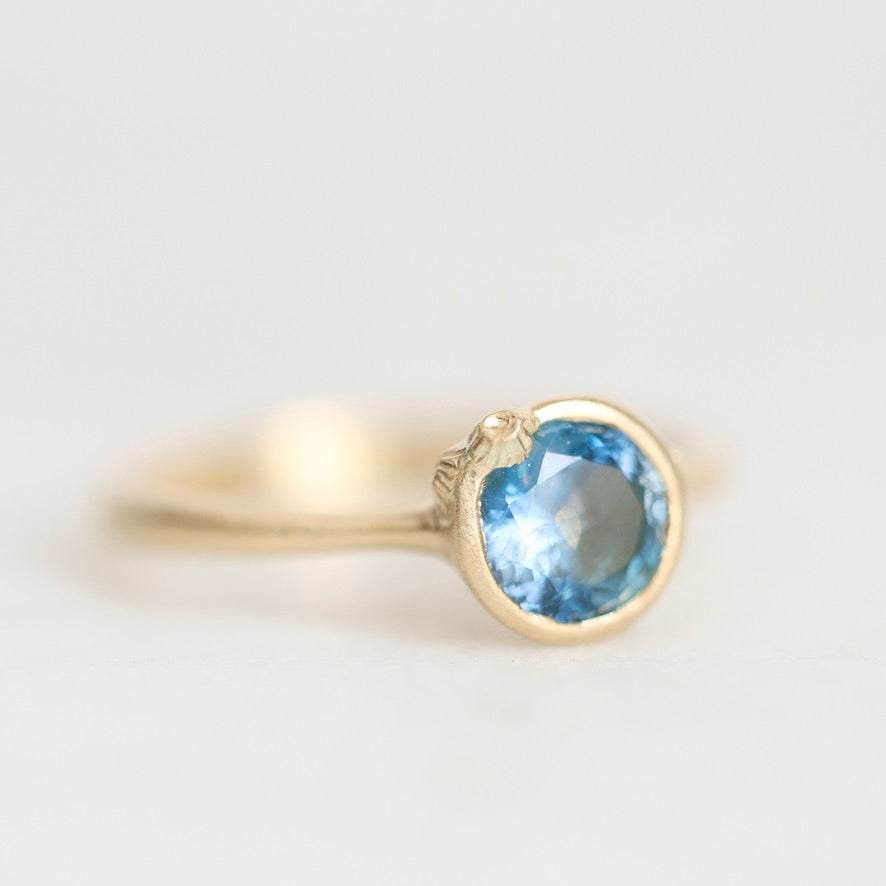 A gold ring with a round-cut light blue gemstone in a bezel setting, photographed on a clean, light gray background.