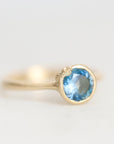 A gold ring with a round-cut light blue gemstone in a bezel setting, photographed on a clean, light gray background.