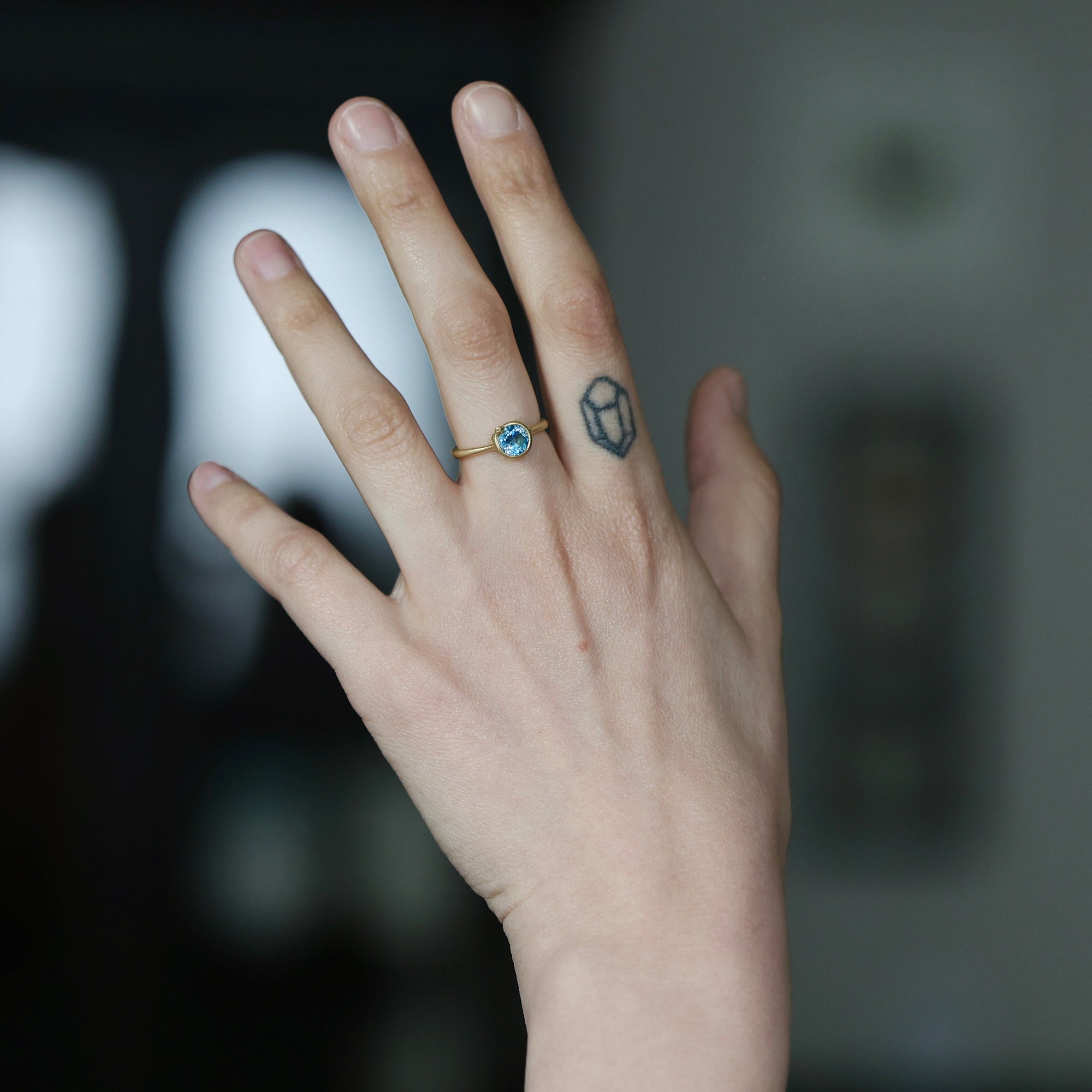 A close-up of a person's hand wearing a delicate gold ring with a small round blue gemstone, set against a dark, blurred background.