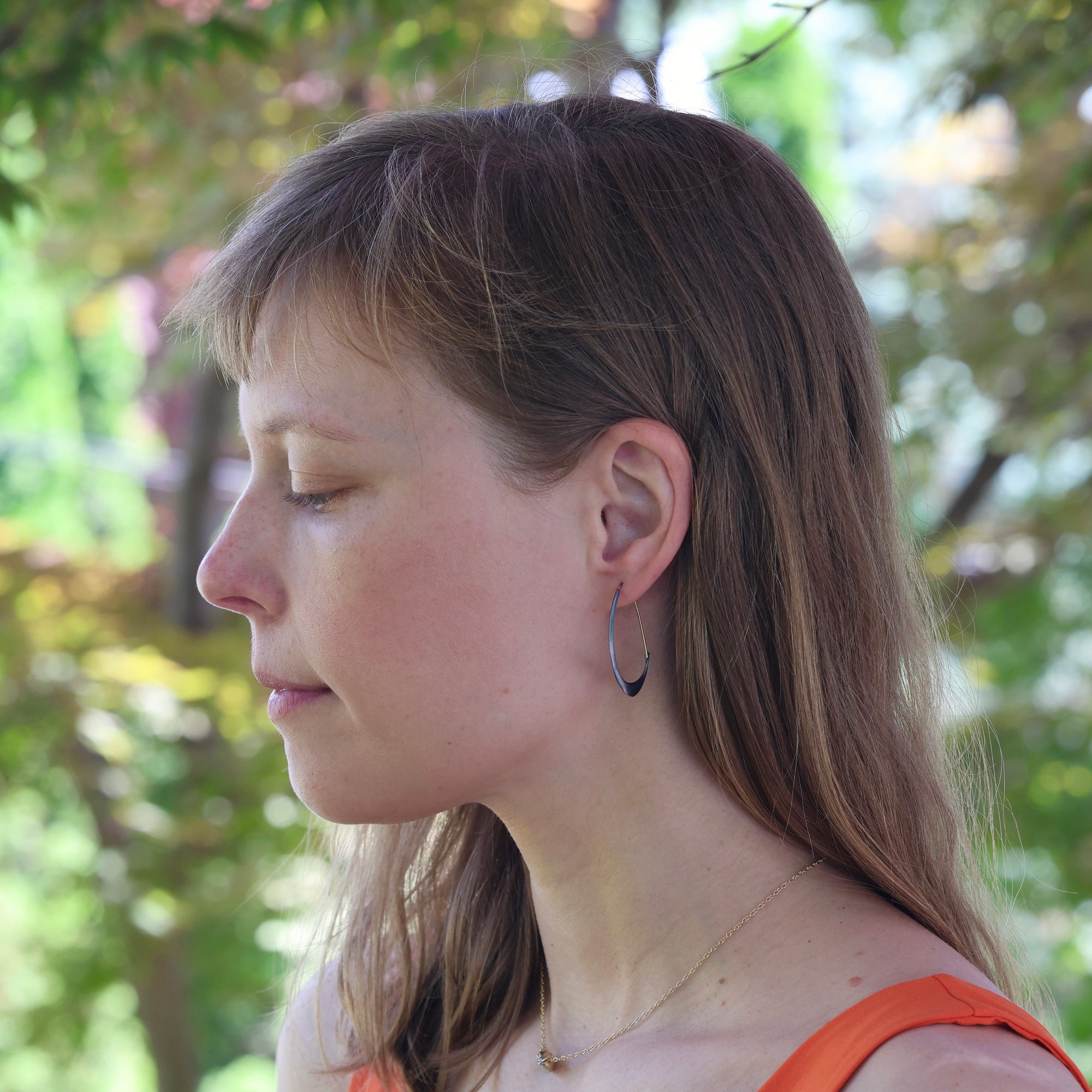 Woman with long brown hair wearing hoop earrings, standing outdoors with greenery in the background