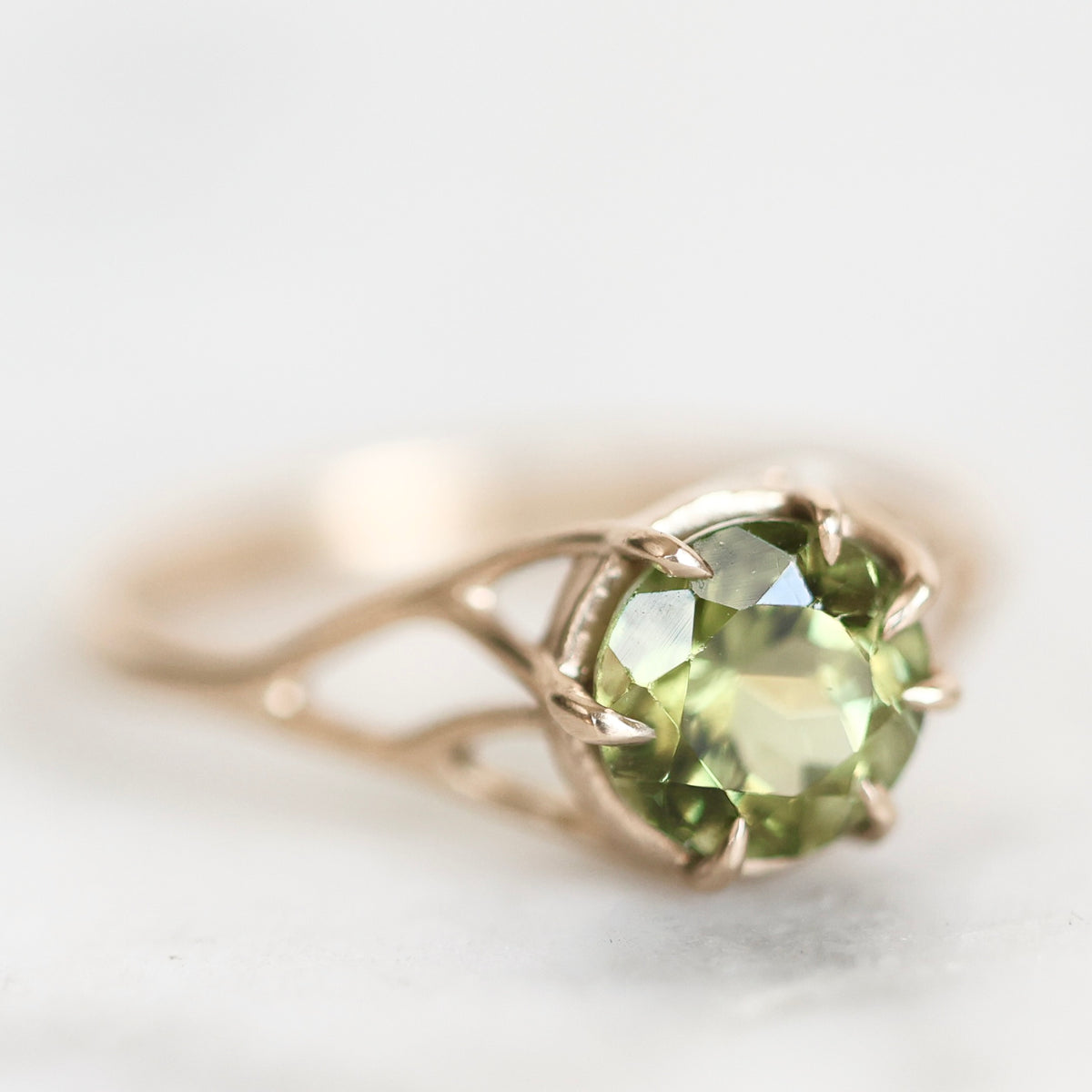 A close-up shot of a rose gold ring featuring a round-cut green peridot gemstone in a prong setting, resting on a light gray surface.