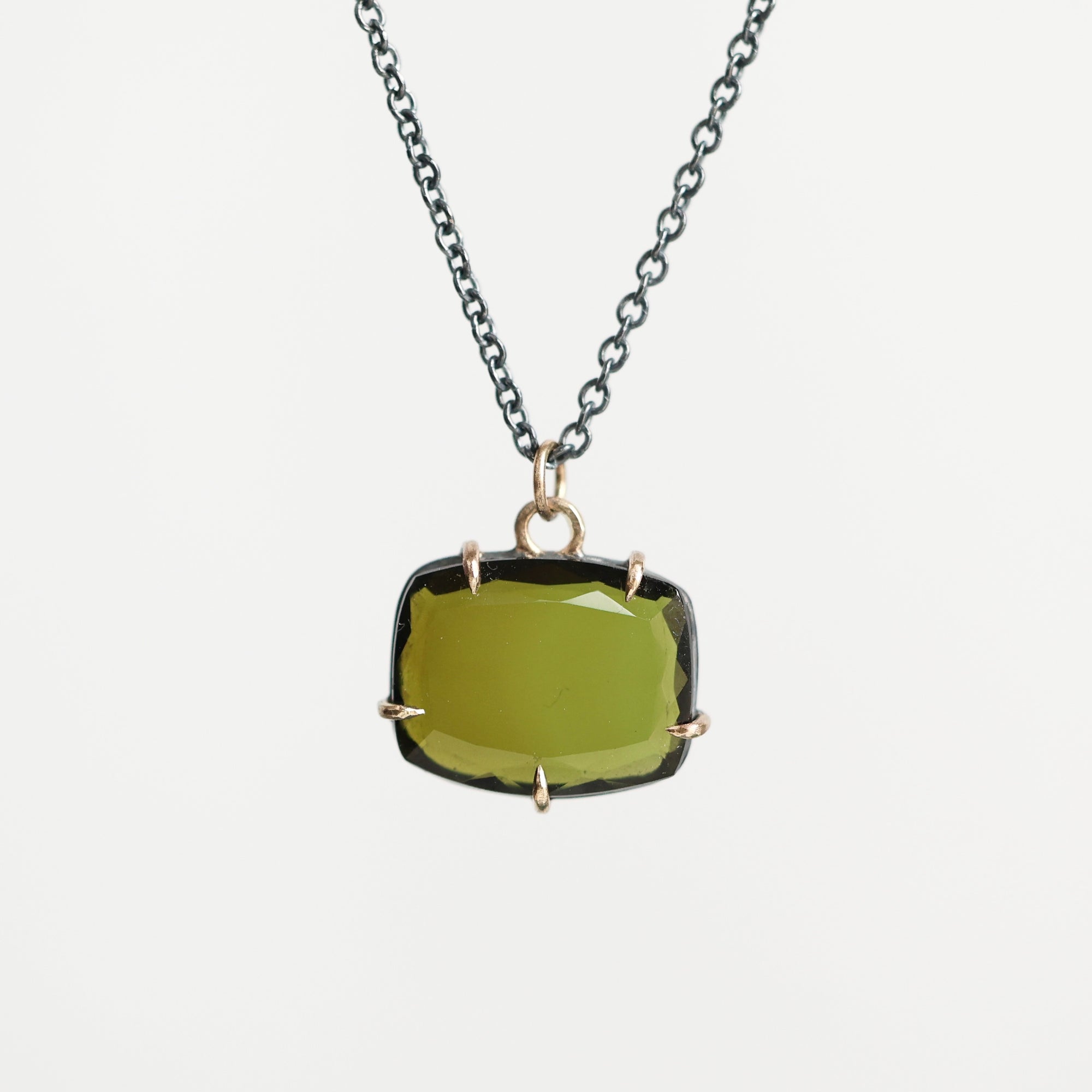 A necklace featuring a large, cushion-cut olive green gemstone in a gold bezel setting, suspended from a dark gunmetal chain against a light gray background.