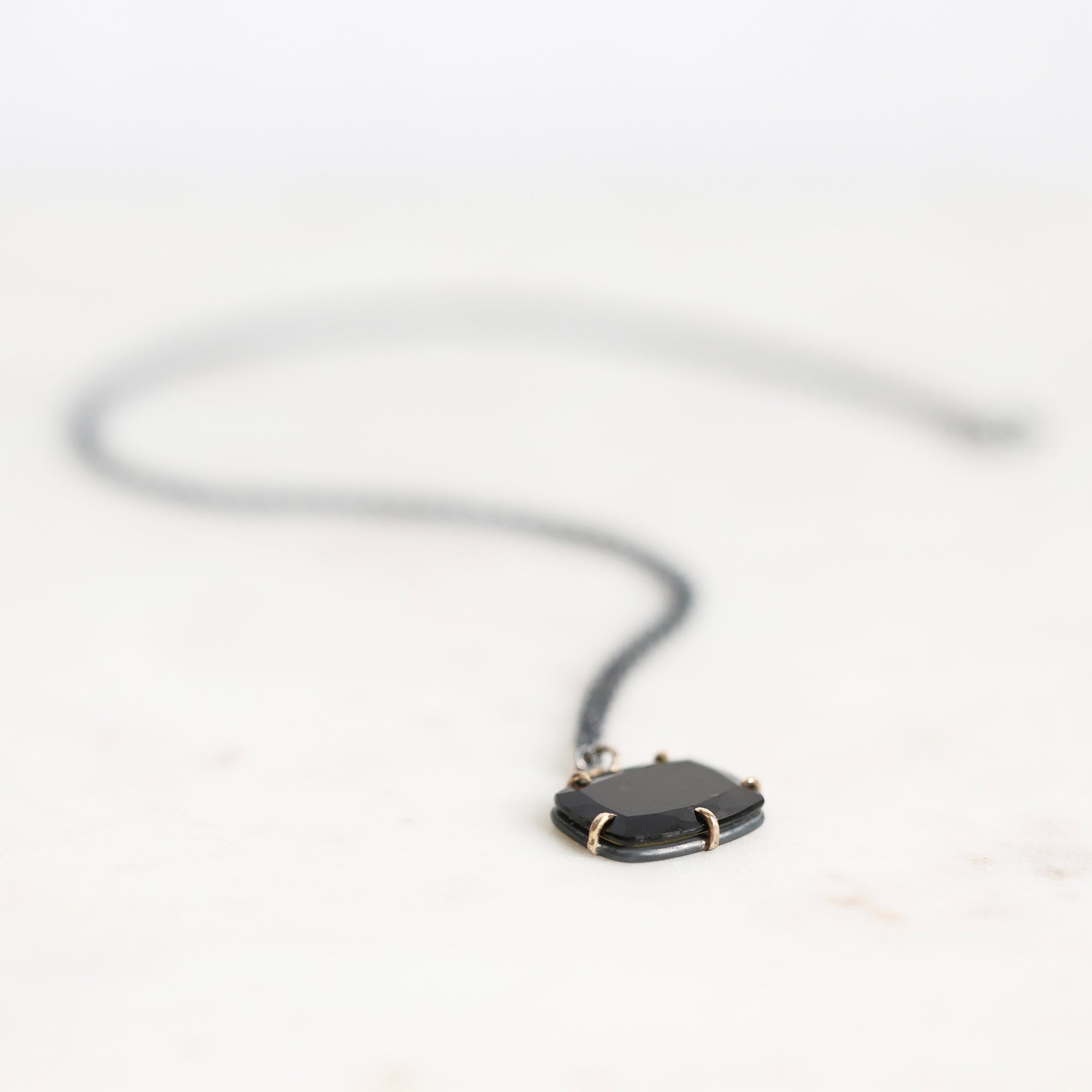 A black pendant necklace with a dark, polished rectangular stone set in a gold-toned bezel, suspended from a thin black cord against a bright white background.