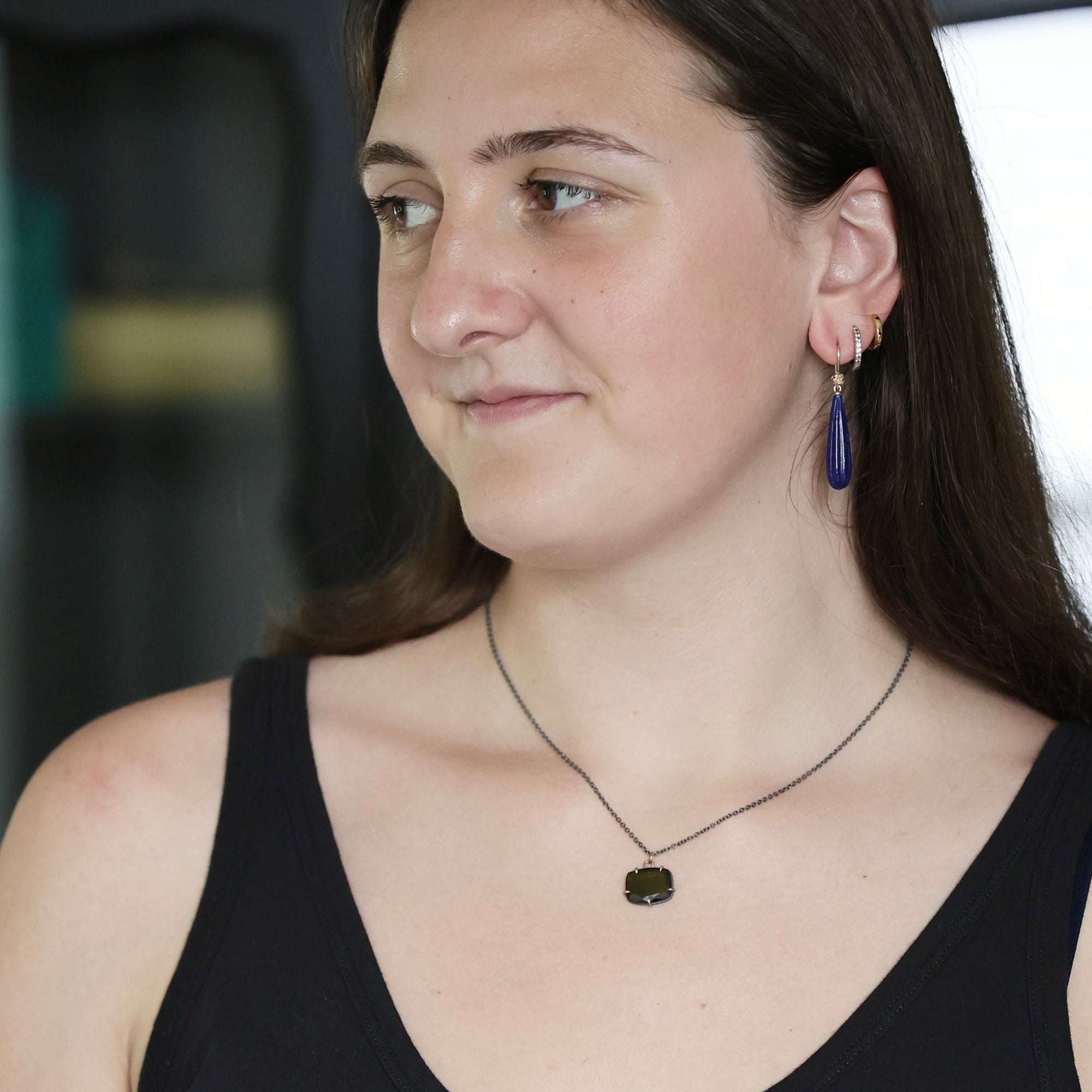 A woman wearing a dark blue teardrop-shaped drop earring and a dark necklace with a small dark pendant, shown in a close-up portrait.