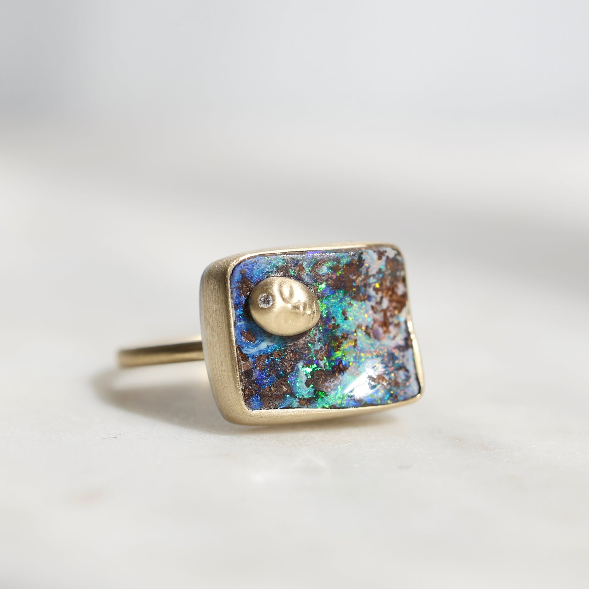 A square bezel-set ring featuring a colorful iridescent opal inlay with blue, green, and brown flashes, accented by a small gold moon and sun motif and a tiny clear stone. The ring is photographed on a light gray surface with a soft, neutral background.