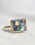 A square bezel-set ring featuring a colorful iridescent opal inlay with blue, green, and brown flashes, accented by a small gold moon and sun motif and a tiny clear stone. The ring is photographed on a light gray surface with a soft, neutral background.