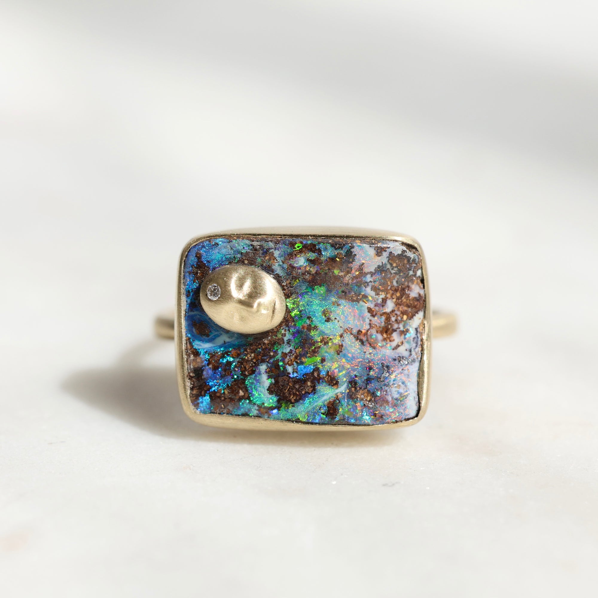 A square bezel-set ring featuring a vibrant blue and brown opal inlay with a small gold crescent moon charm and a tiny clear stone, shown on a light neutral background.