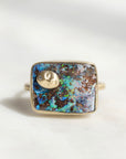 A square bezel-set ring featuring a vibrant blue and brown opal inlay with a small gold crescent moon charm and a tiny clear stone, shown on a light neutral background.