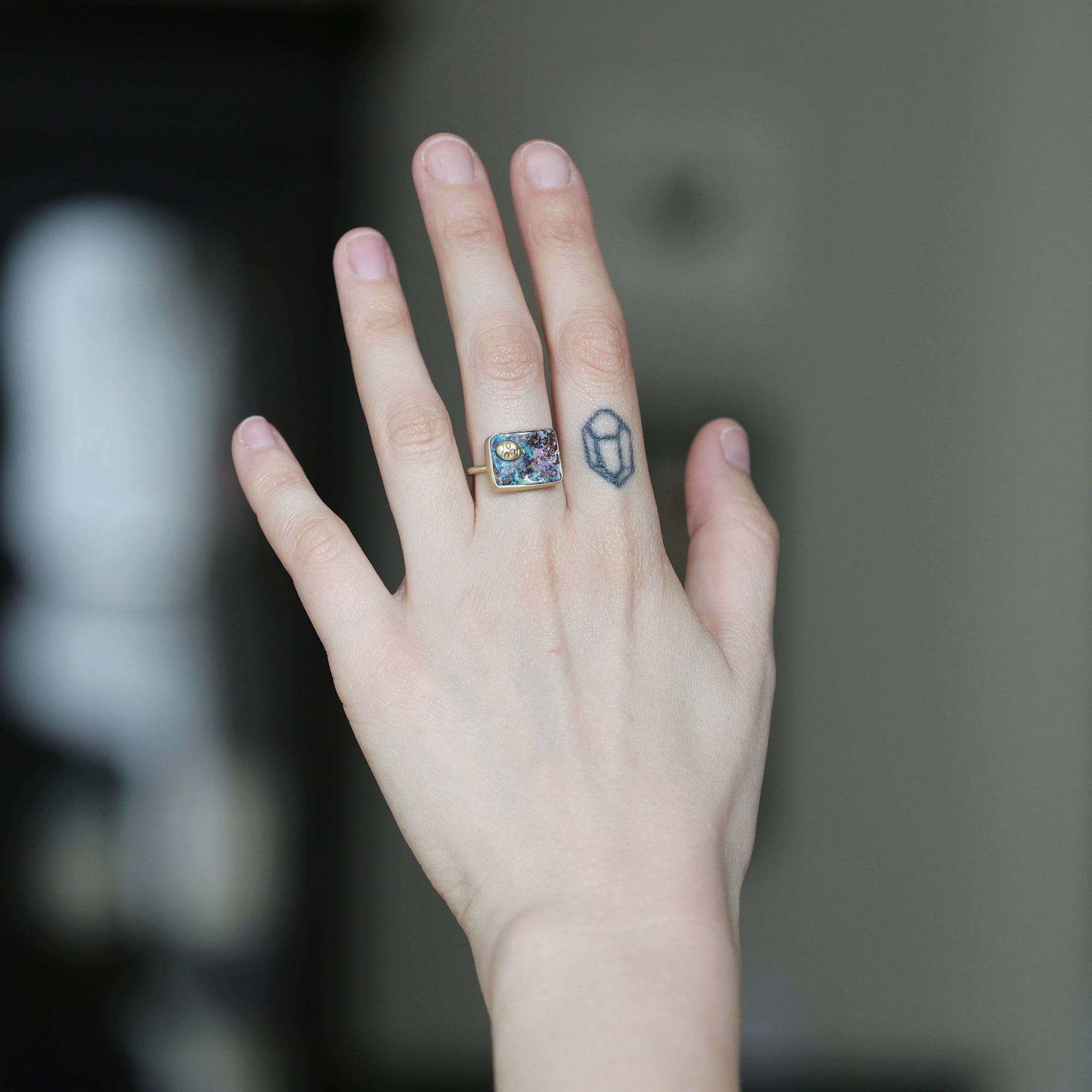 A close-up of a person's hand wearing a gold ring with a colorful, textured square face and a small geometric tattoo on the side of the hand.