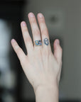 A close-up of a person's hand wearing a gold ring with a colorful, textured square face and a small geometric tattoo on the side of the hand.