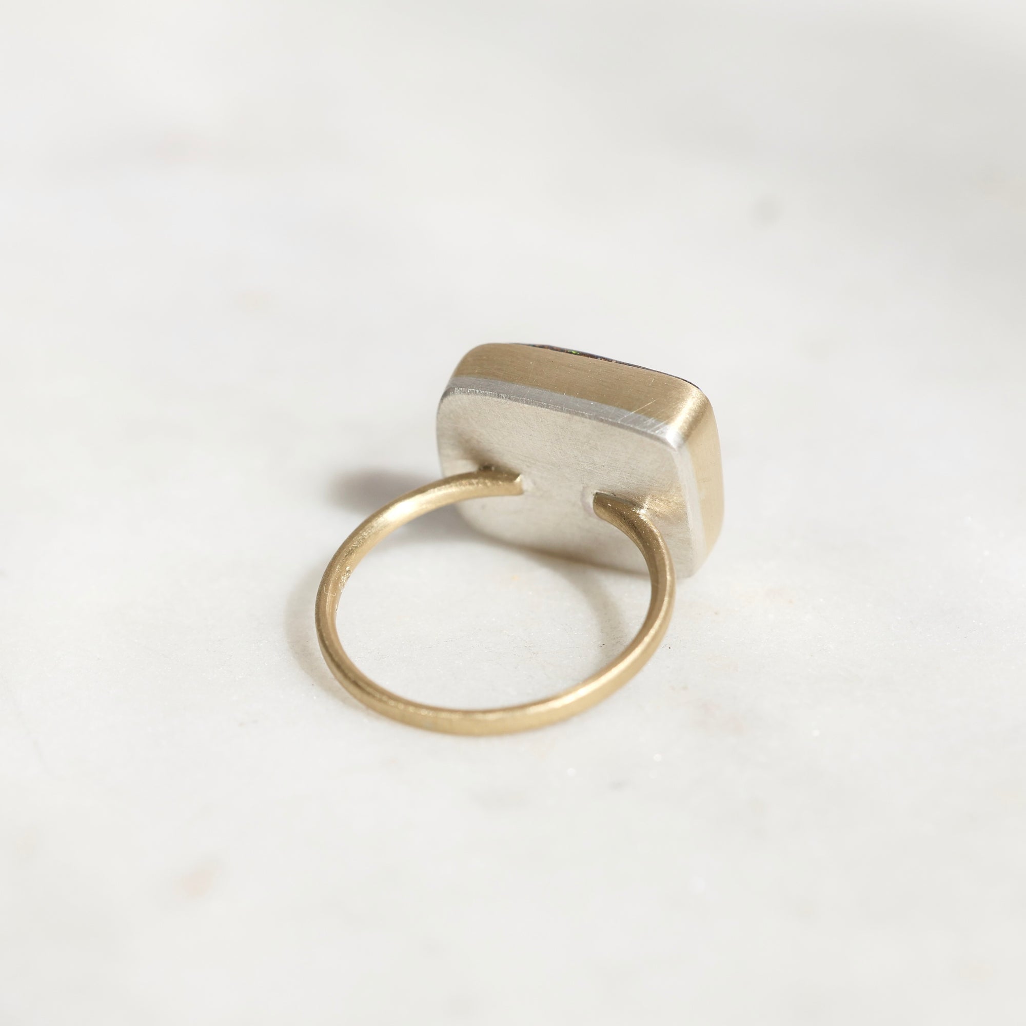 A minimalist ring featuring a square, beveled silver-toned face set on a thin, polished gold band, photographed on a light, neutral marble surface.