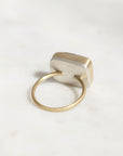 A minimalist ring featuring a square, beveled silver-toned face set on a thin, polished gold band, photographed on a light, neutral marble surface.