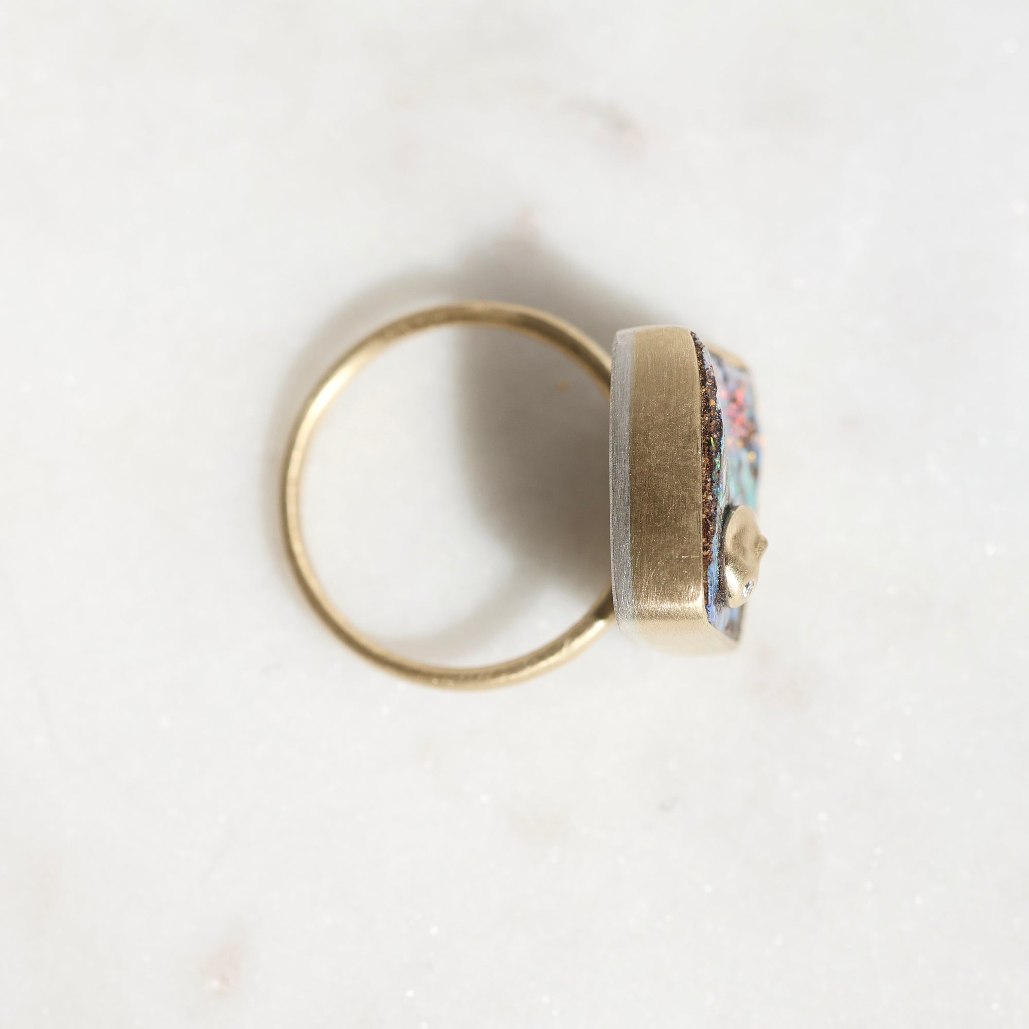 A gold ring with a square face featuring a colorful floral enamel inlay and a small gold heart, shown on a light marble surface.
