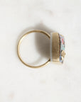 A gold ring with a square face featuring a colorful floral enamel inlay and a small gold heart, shown on a light marble surface.