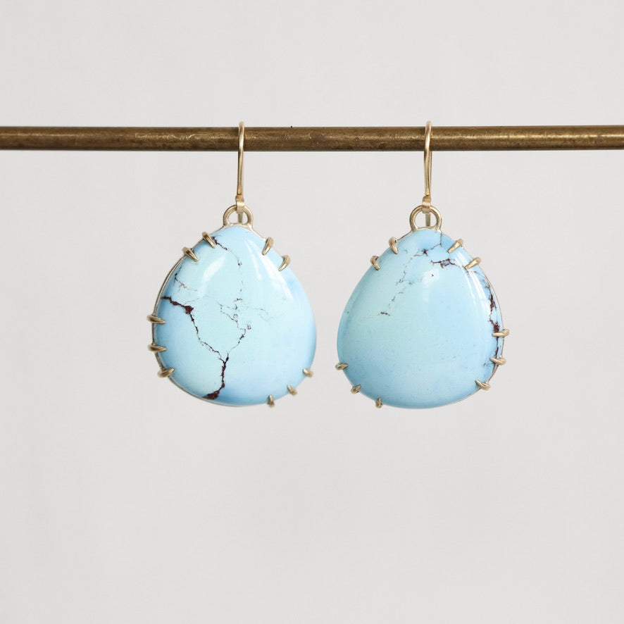 A pair of teardrop-shaped light blue turquoise drop earrings with gold hooks and gold bezel settings, hanging from a gold horizontal bar against a light gray background.