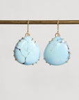 A pair of teardrop-shaped light blue turquoise drop earrings with gold hooks and gold bezel settings, hanging from a gold horizontal bar against a light gray background.