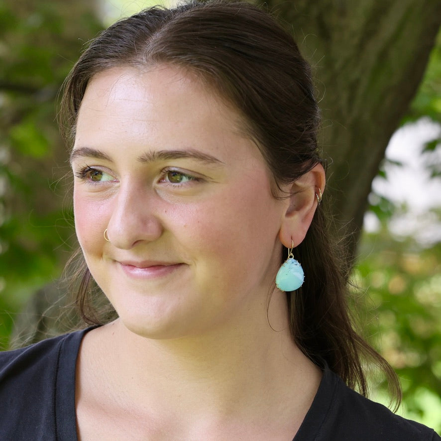 A woman outdoors wearing a light blue teardrop-shaped dangle earring with a gold hoop, shown in a close-up portrait.