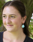 A woman outdoors wearing a light blue teardrop-shaped dangle earring with a gold hoop, shown in a close-up portrait.