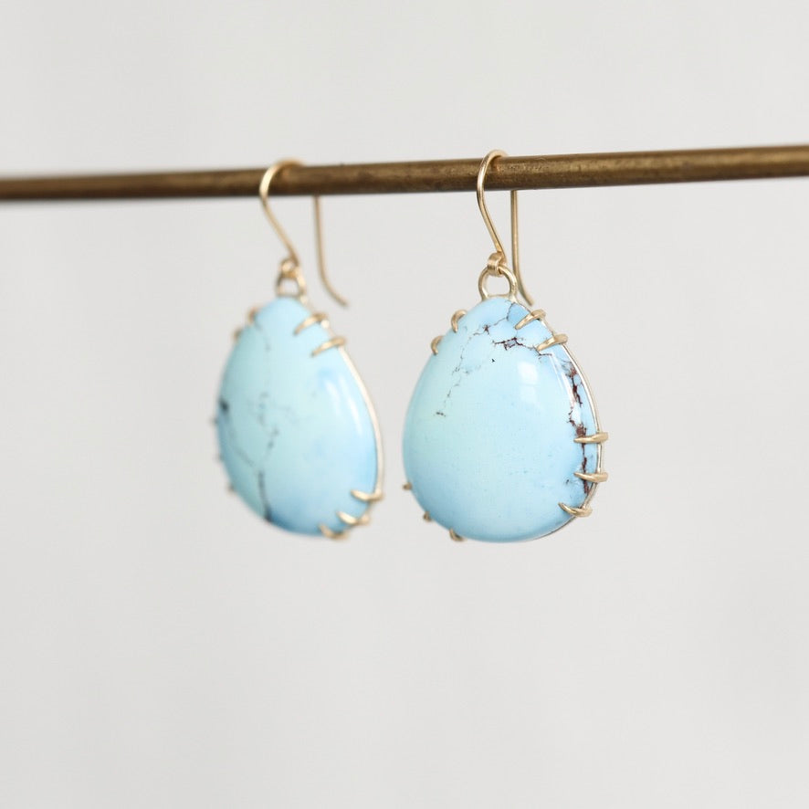 A pair of teardrop-shaped light blue turquoise drop earrings with gold wire-wrapped edges and gold fishhook wires, hanging from a gold-colored metal rod against a plain light gray background.