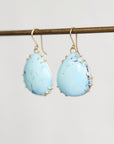 A pair of teardrop-shaped light blue turquoise drop earrings with gold wire-wrapped edges and gold fishhook wires, hanging from a gold-colored metal rod against a plain light gray background.