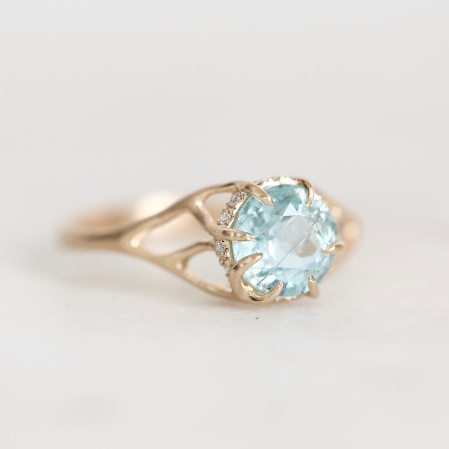 A yellow gold ring featuring a round-cut light blue aquamarine gemstone in a prong setting, accented by small clear stones along the band, shown on a plain light gray background.