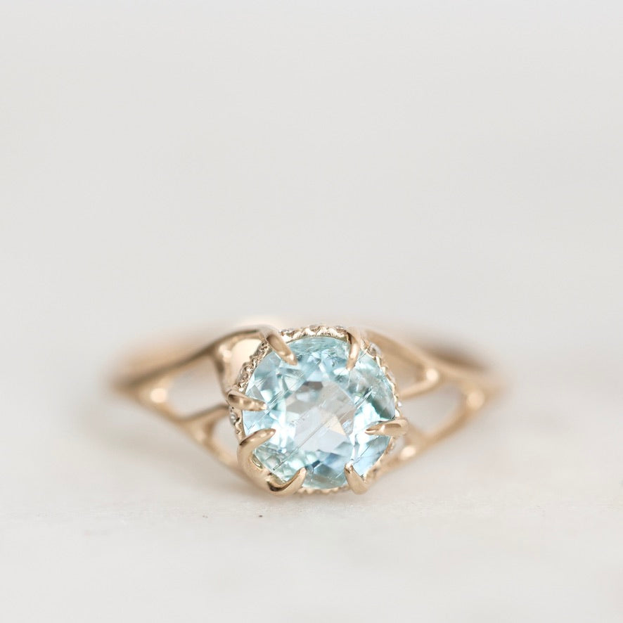 A gold ring featuring a large, round-cut light blue gemstone in a prong setting, shown on a soft, light gray background.