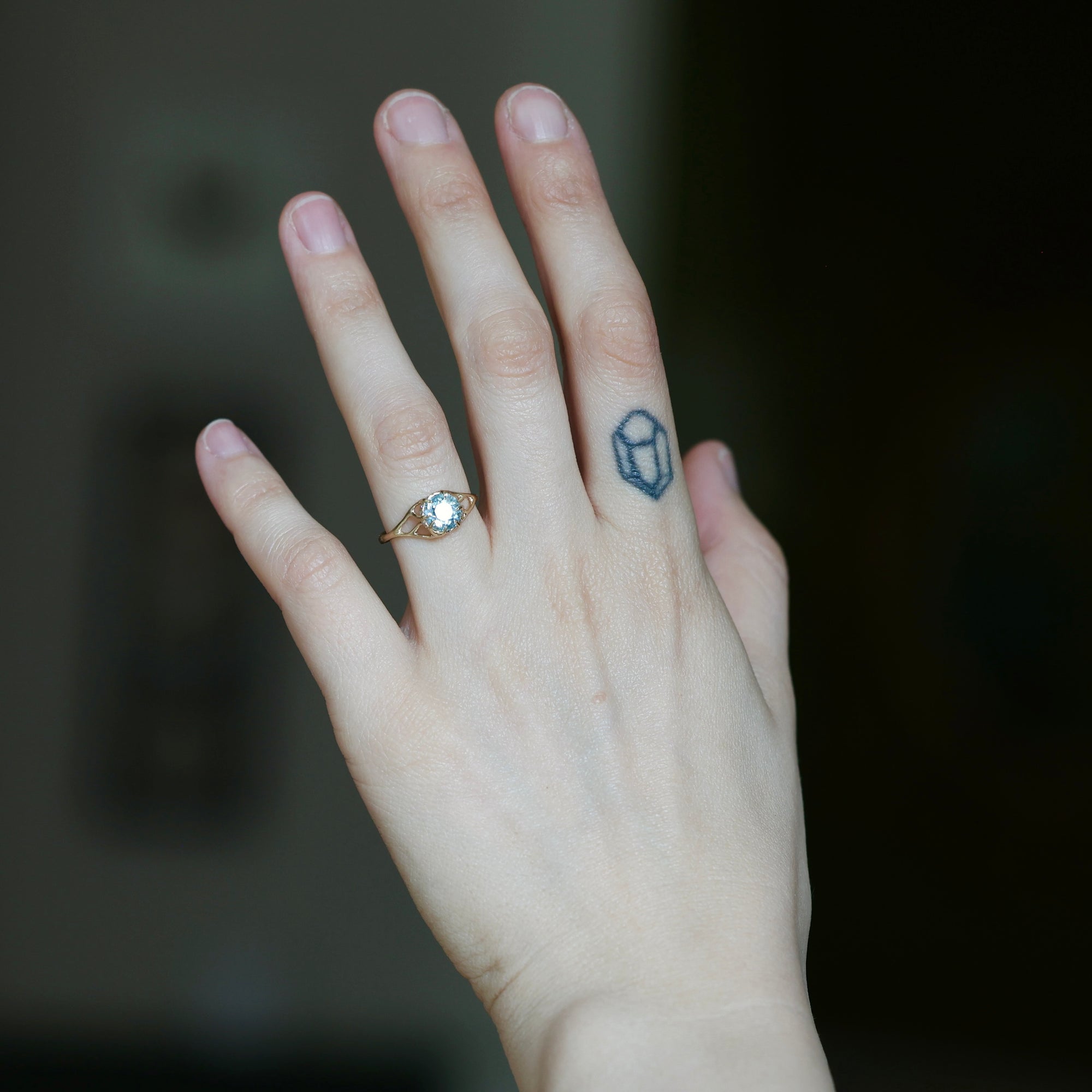 A close-up of a person's hand wearing a gold ring with a clear round gemstone on the ring finger, with a small blue geometric tattoo on the middle finger, against a dark blurred background.