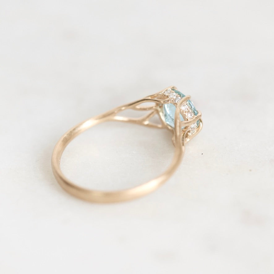 A yellow gold ring featuring a light blue gemstone and small clear accent stones, photographed on a light gray background.