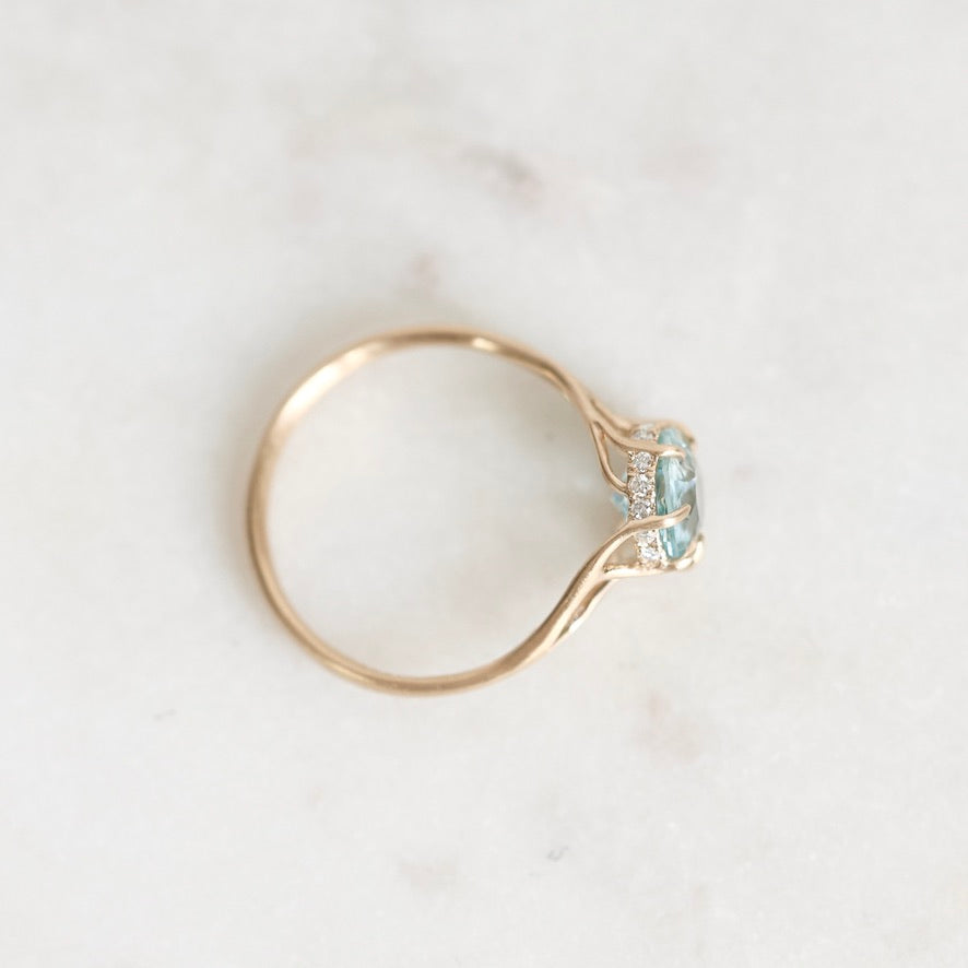 A gold ring with a small gemstone and tiny clear accent stones, photographed on a pale marble-like surface.