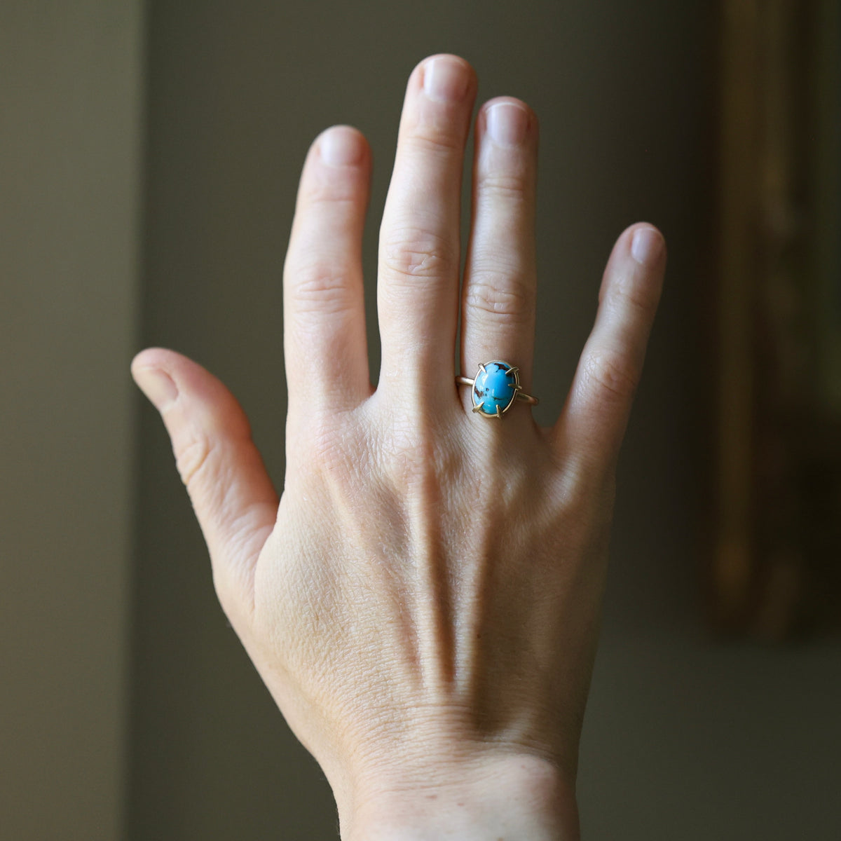 A hand wearing a gold ring with a blue-green cabochon stone, shown against a soft-focus indoor background.