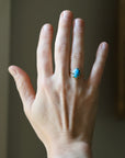 A hand wearing a gold ring with a blue-green cabochon stone, shown against a soft-focus indoor background.