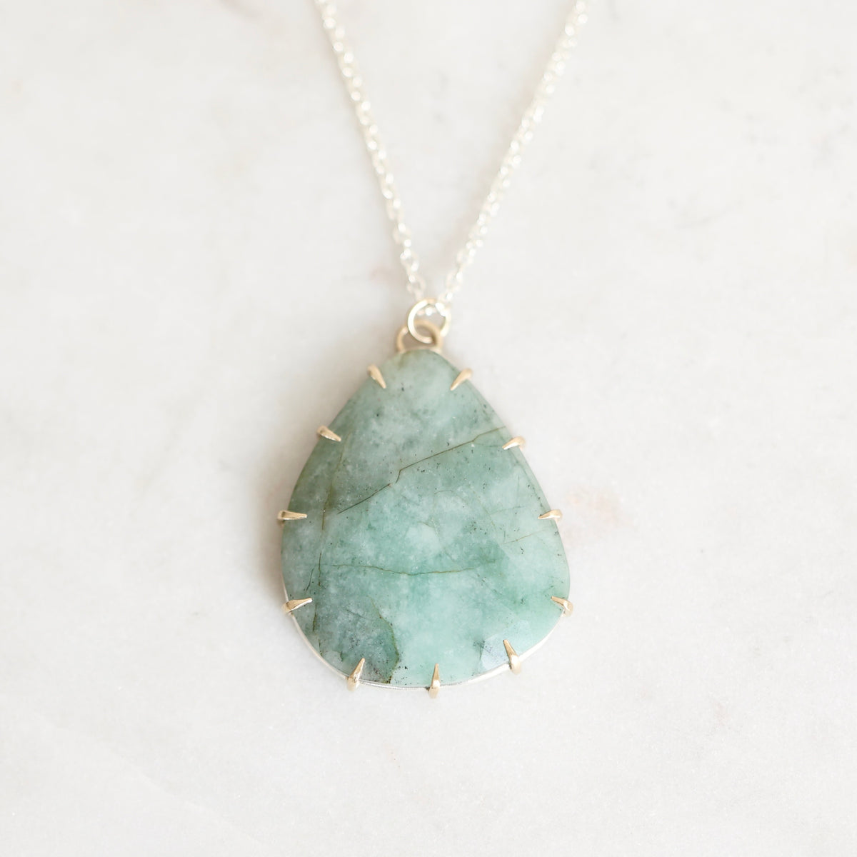 A teardrop-shaped green stone pendant necklace with a gold prongs and small gold spikes around the edges, hanging from a delicate gold chain on a light marble background.