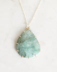 A teardrop-shaped green stone pendant necklace with a gold prongs and small gold spikes around the edges, hanging from a delicate gold chain on a light marble background.