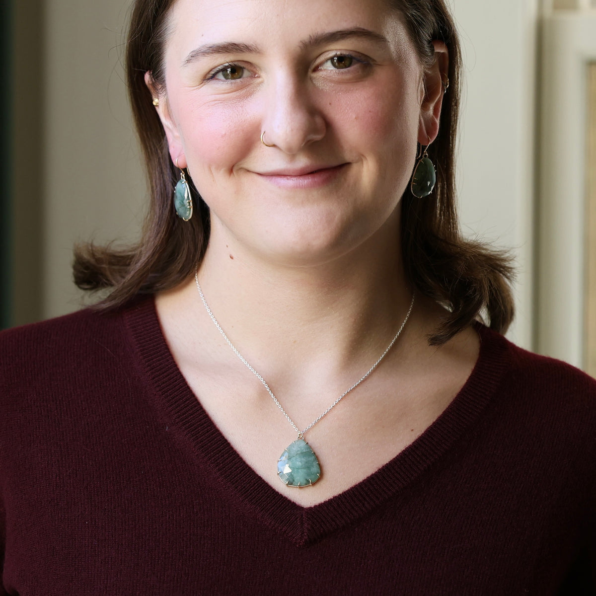 A woman wearing a silver pendant necklace with a green teardrop-shaped stone and matching green teardrop earrings, paired with a burgundy V-neck sweater.