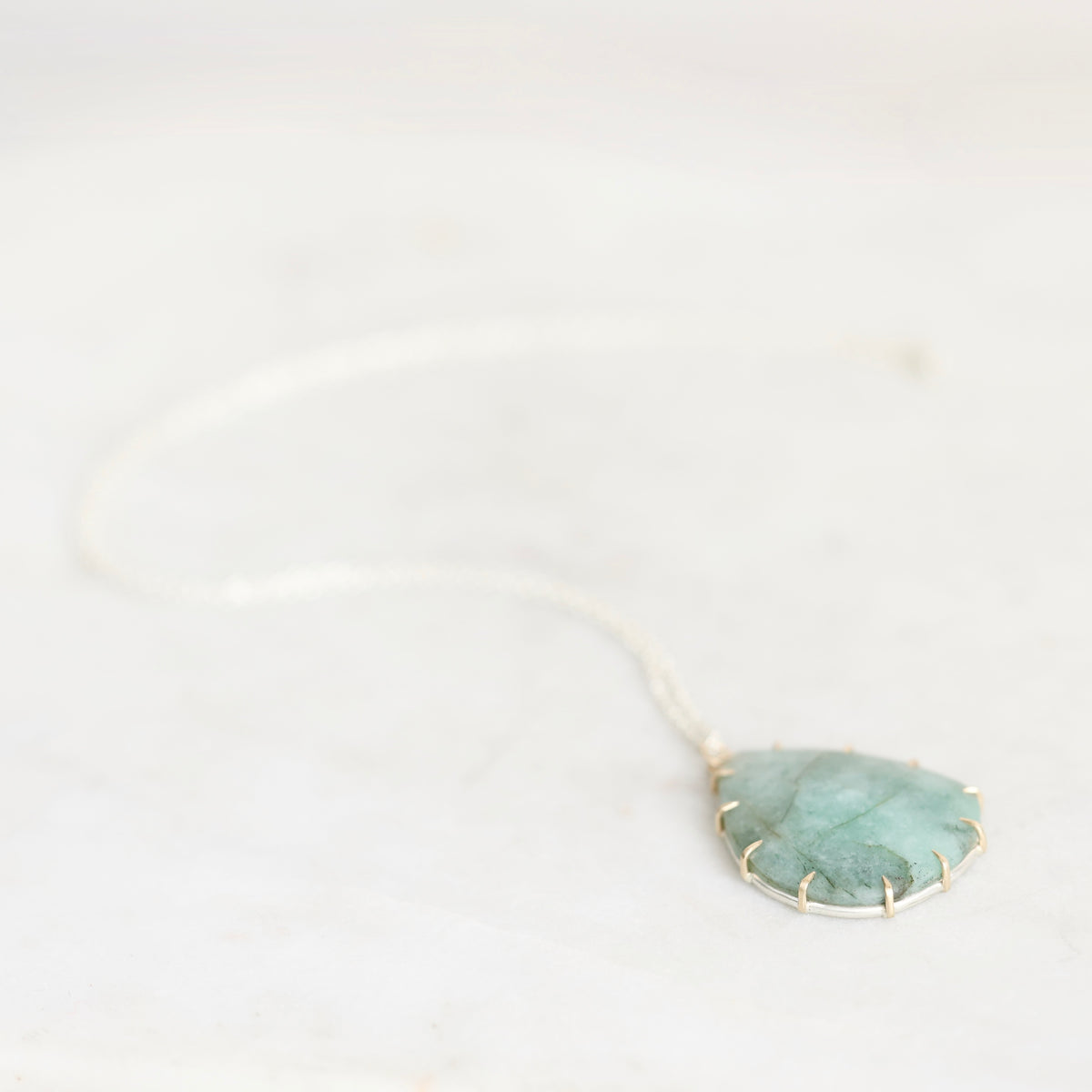 A delicate silver chain necklace featuring a leaf-shaped pendant made of textured green stone, held in a gold bezel setting, resting on a light marble surface.