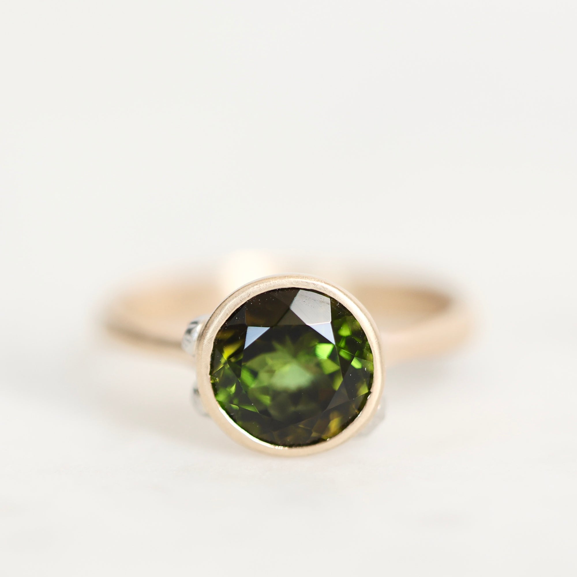 A round bezel-set gemstone ring featuring a faceted olive green stone in a gold-toned setting, photographed on a clean white background.