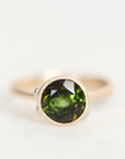 A round bezel-set gemstone ring featuring a faceted olive green stone in a gold-toned setting, photographed on a clean white background.