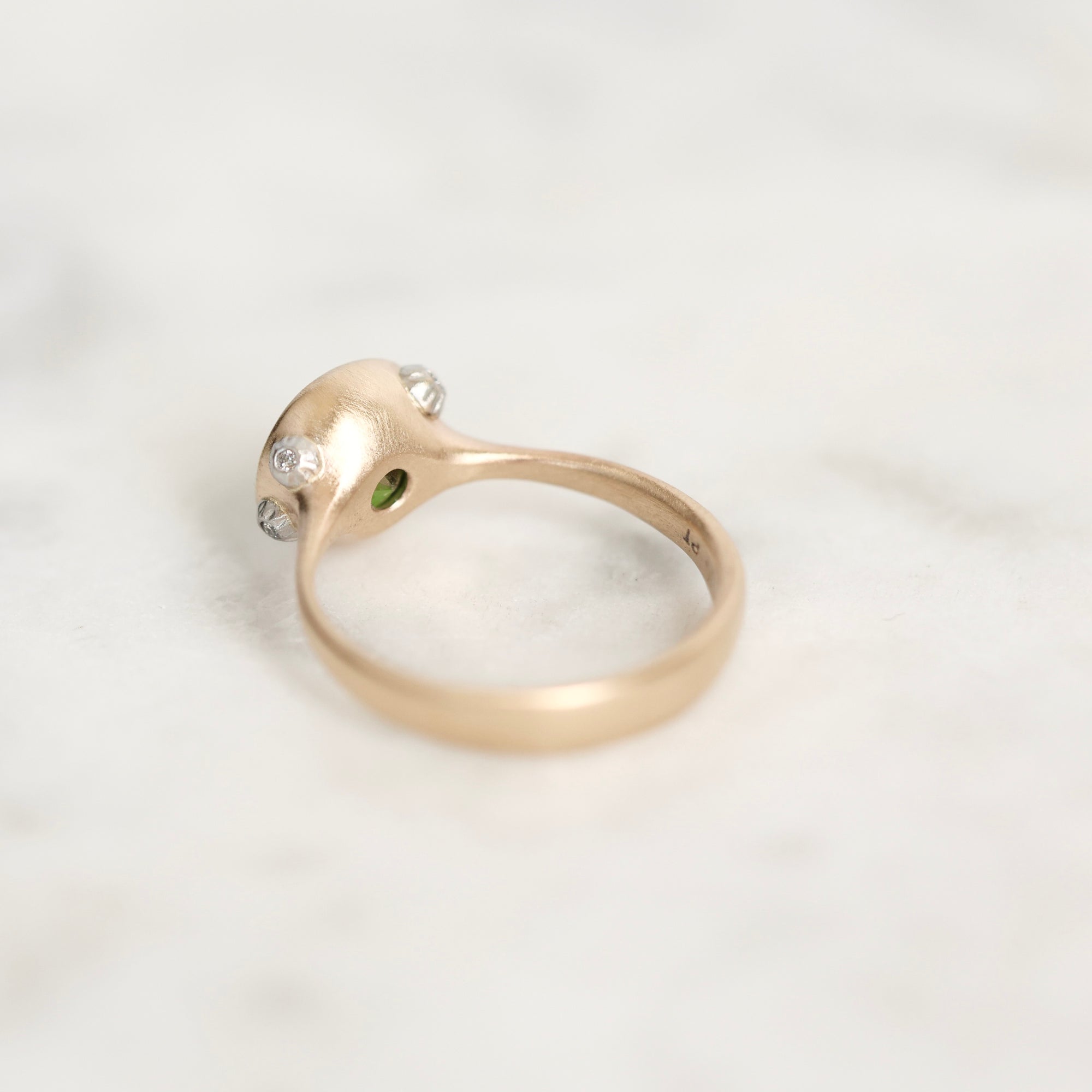A gold ring with a sculpted dome-shaped top set with small clear stones and a tiny green accent, resting on a light marble surface.