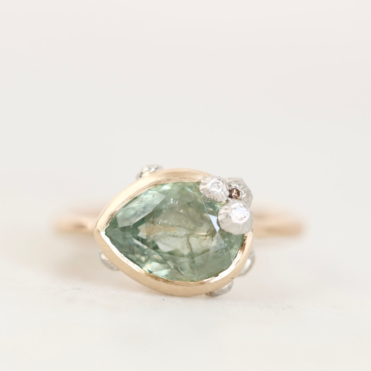 A vintage-style ring featuring a large pear-cut light green gemstone in a gold setting, accented with small clear stones on the right side, photographed against a plain off-white background.
