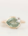 A vintage-style ring featuring a large pear-cut light green gemstone in a gold setting, accented with small clear stones on the right side, photographed against a plain off-white background.