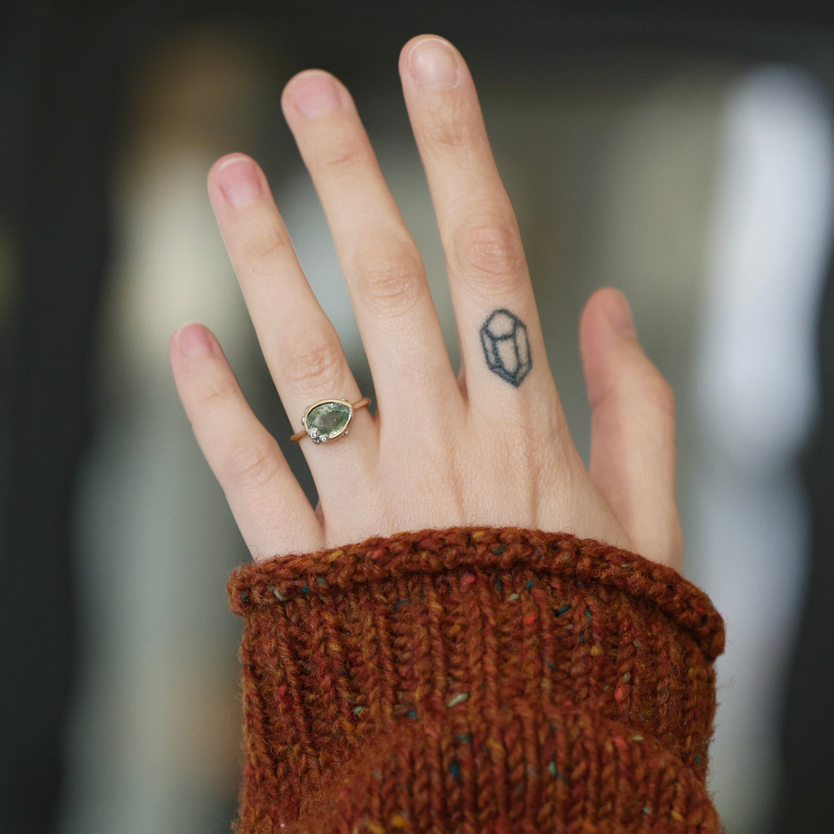 A close-up of a person's hand wearing a gold ring with a green gemstone, styled with a dark geometric tattoo on the middle finger and a rust-colored knit sweater cuff.