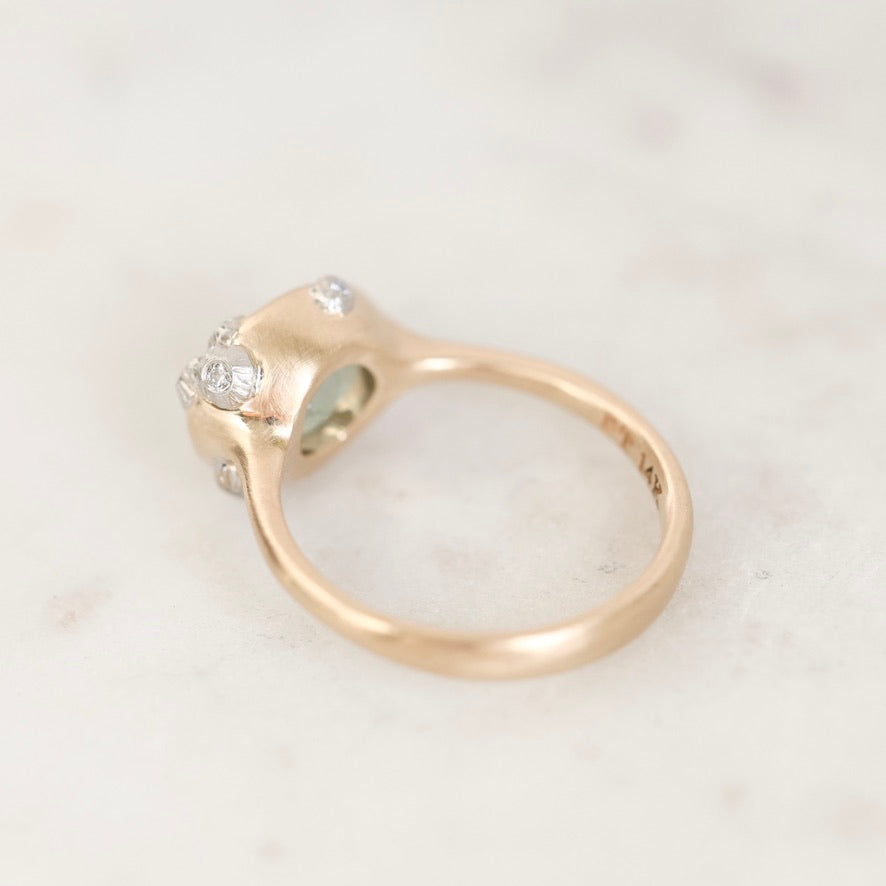 A close-up shot of a yellow gold ring with a cluster of small clear stones on a light marble surface.